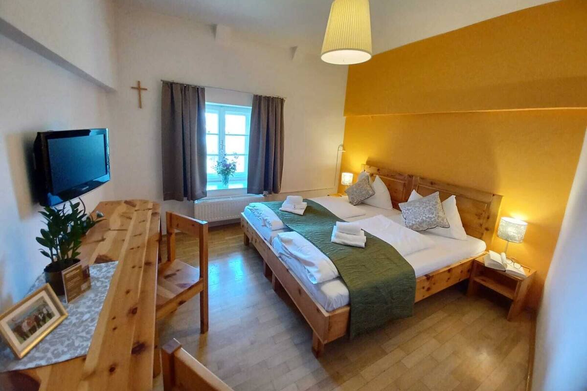 Quarto de hotel com cama, mesa de cabeceira, luminária, televisão, janela com cortinas e uma cruz na parede.