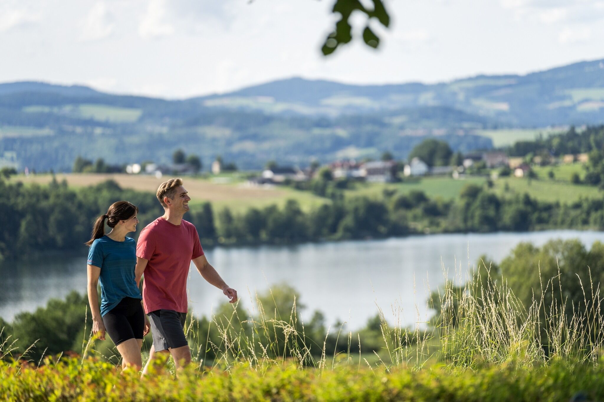 Ett par promenerar på en gräsklädd kulle med utsikt över en sjö, och i fjärran syns hus.