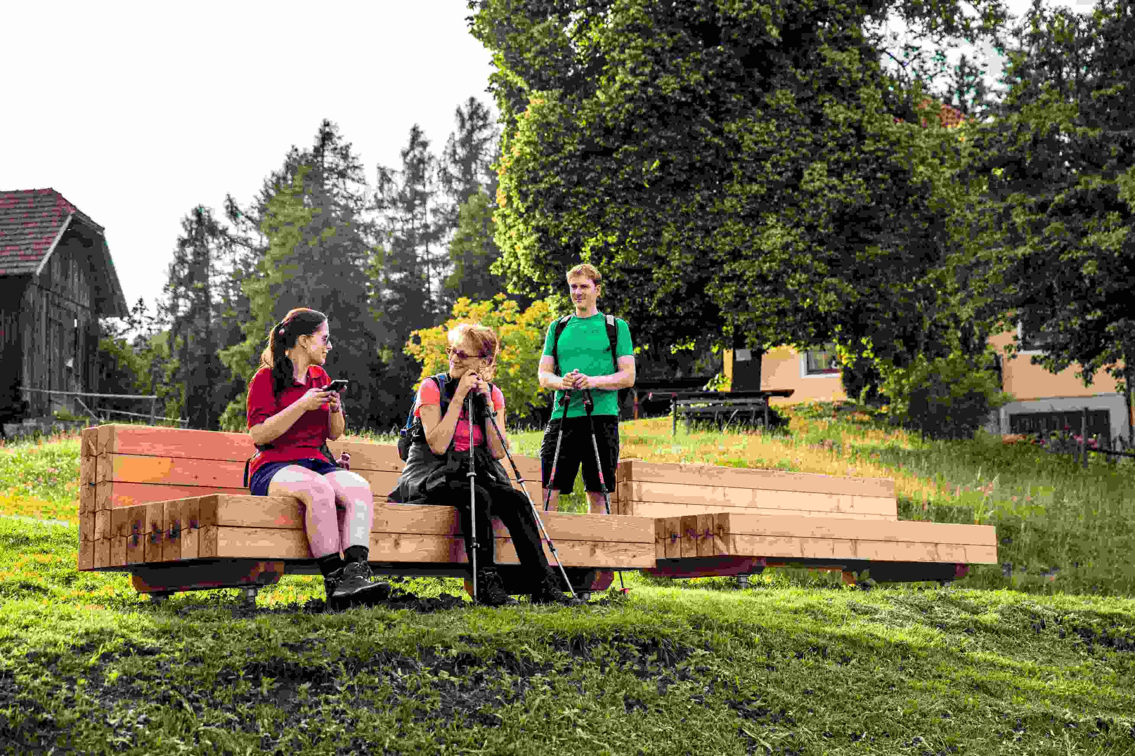 A group of people sitting on a wooden bench in a grassy area with trees and a building in the background.