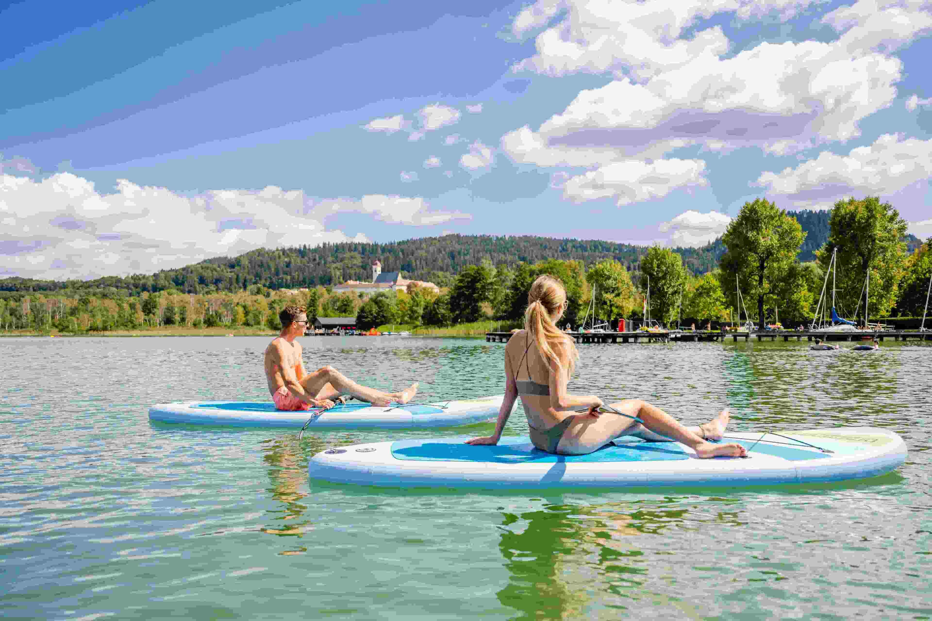 Two individuals sitting on paddleboards on a calm lake with a forested hill in the background.