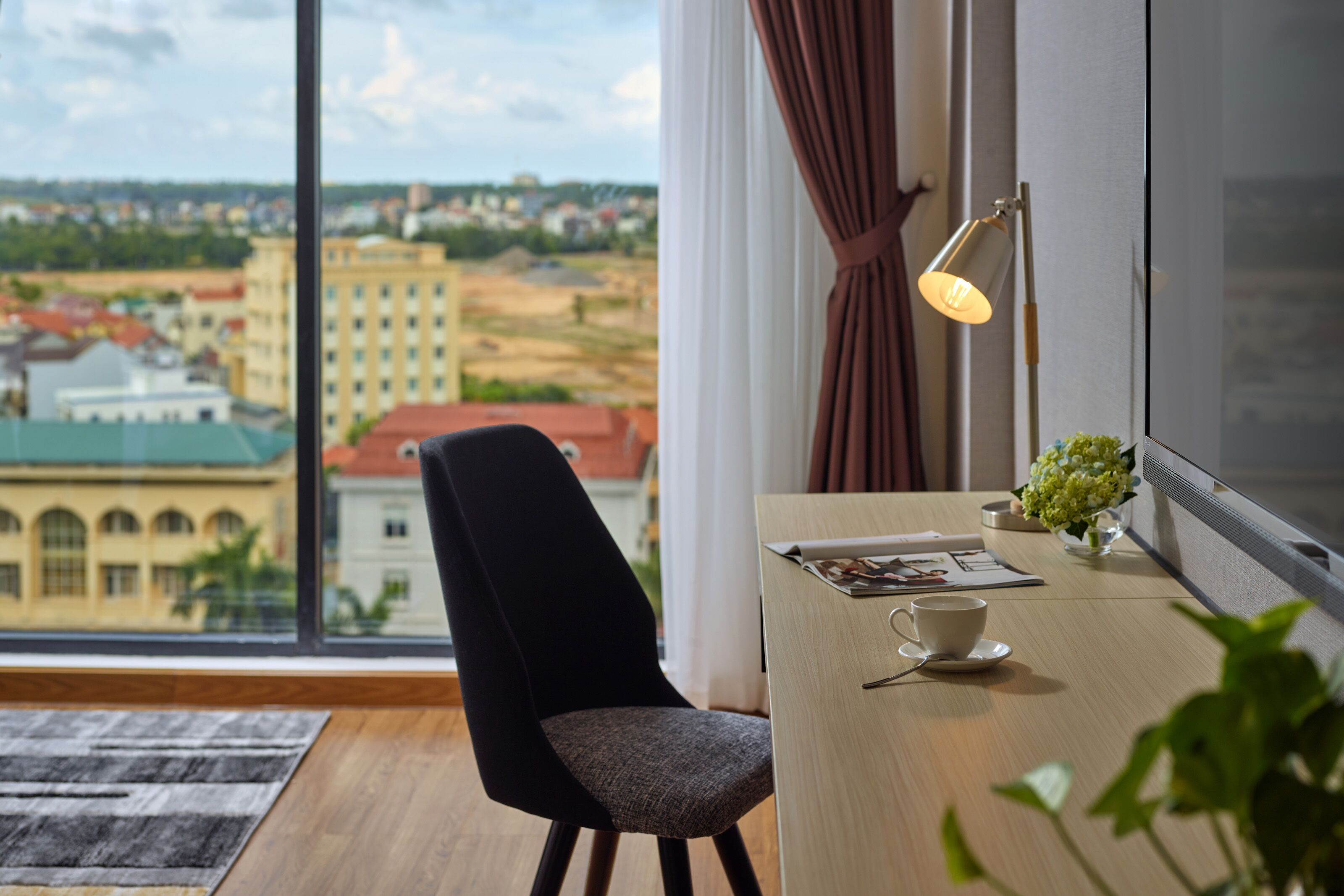 Een moderne kamer met een groot raam, een bureau met kop en schotel, een lamp en een stoel.