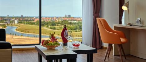 Una habitación moderna con un ventanal amplio que ofrece vistas a un río y edificios lejanos. Hay una mesa con fruteros, una copa de vino y una botella. Se ve una silla naranja y un escritorio con una lámpara.