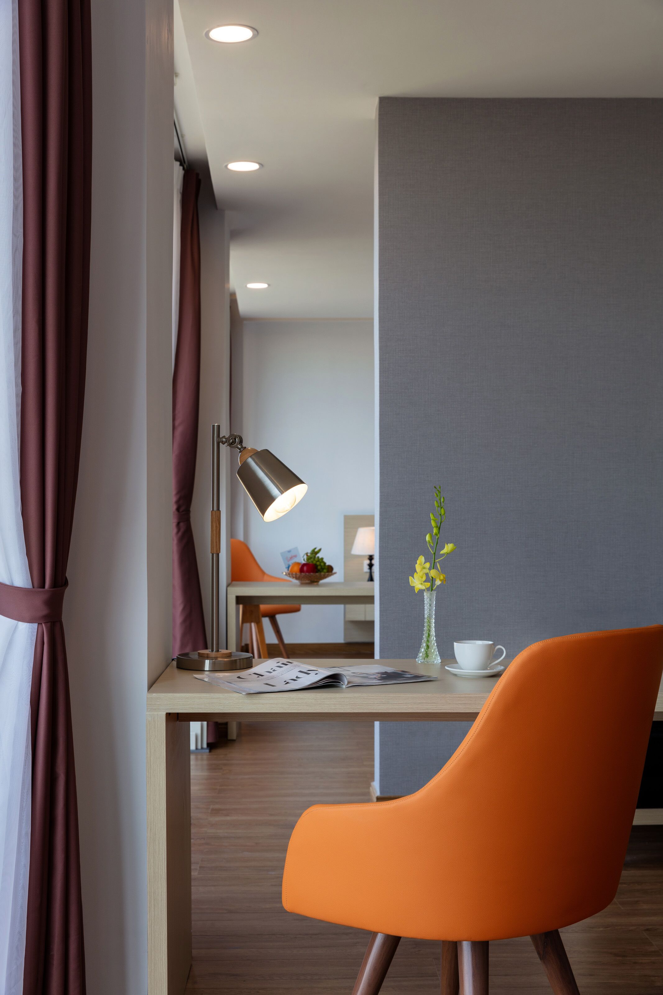 Una habitación de hotel moderna con un escritorio de madera, una silla naranja, una lámpara de escritorio, un jarrón con flores y vistas a un paisaje urbano a través de amplias ventanas.