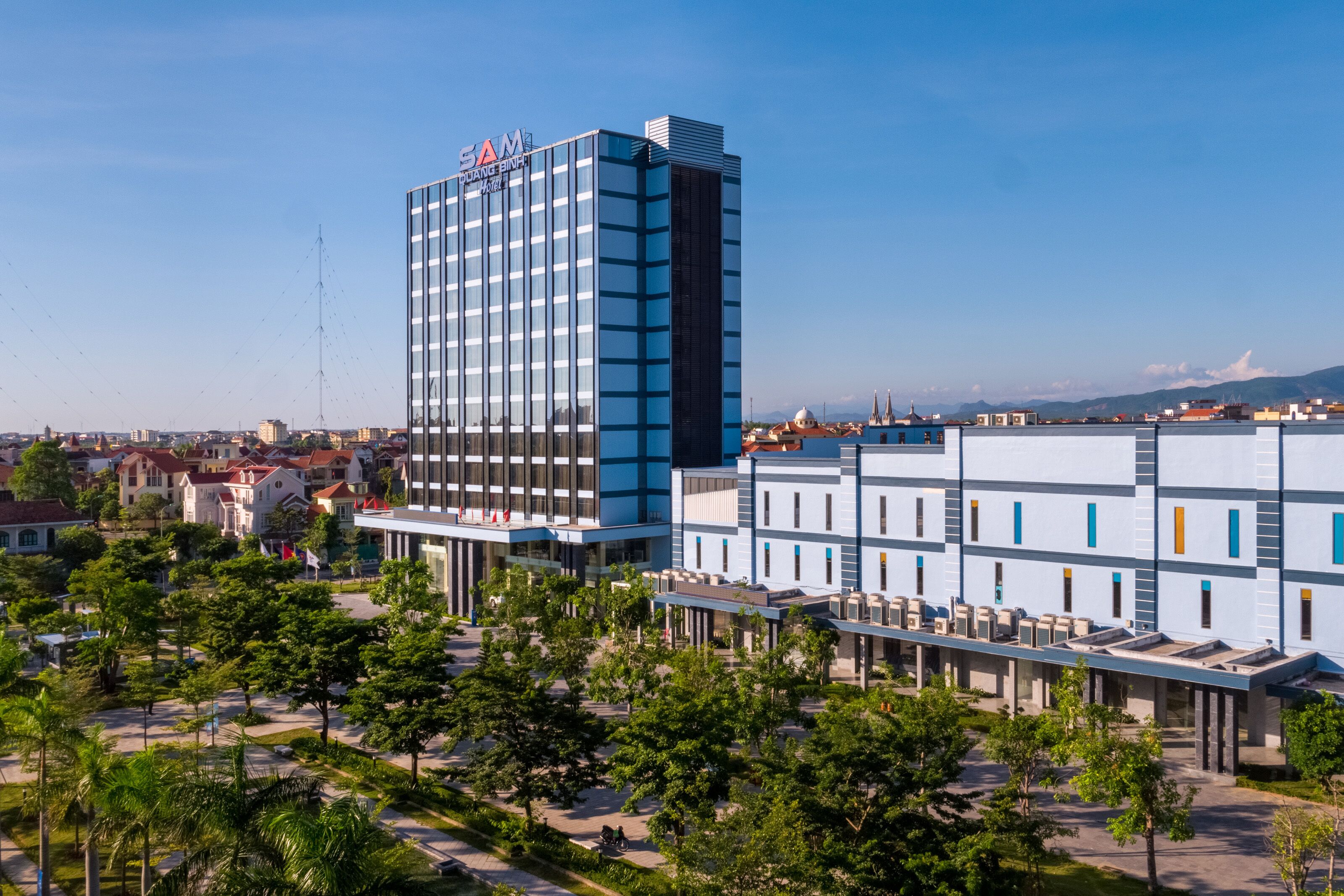 Um hotel moderno com fachada de vidro destacada, um pátio central com árvores e vista para a paisagem urbana ao redor.