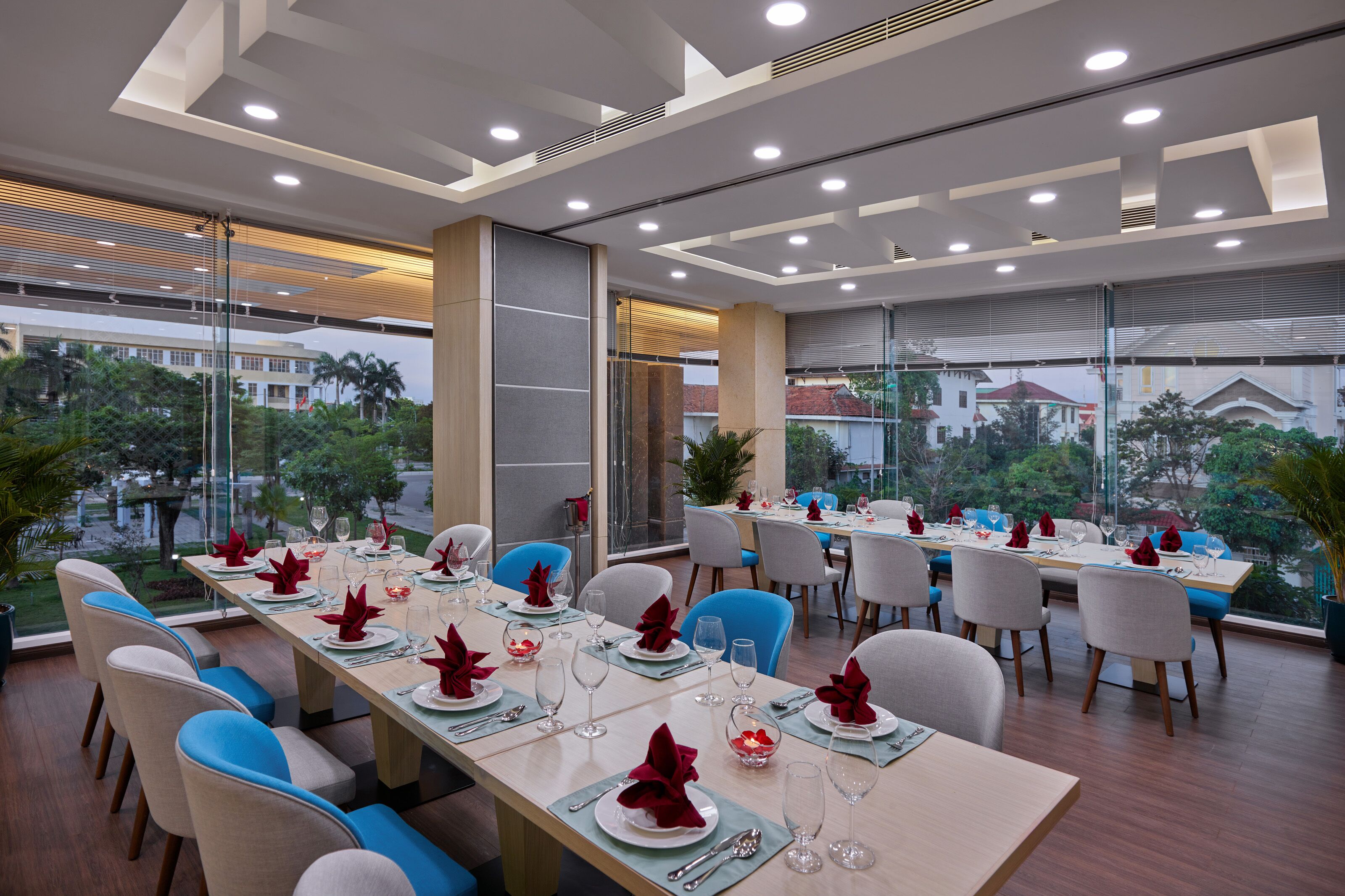 Uma sala de jantar com mesas preparadas para refeição, grandes janelas e vista para edifícios e árvores.