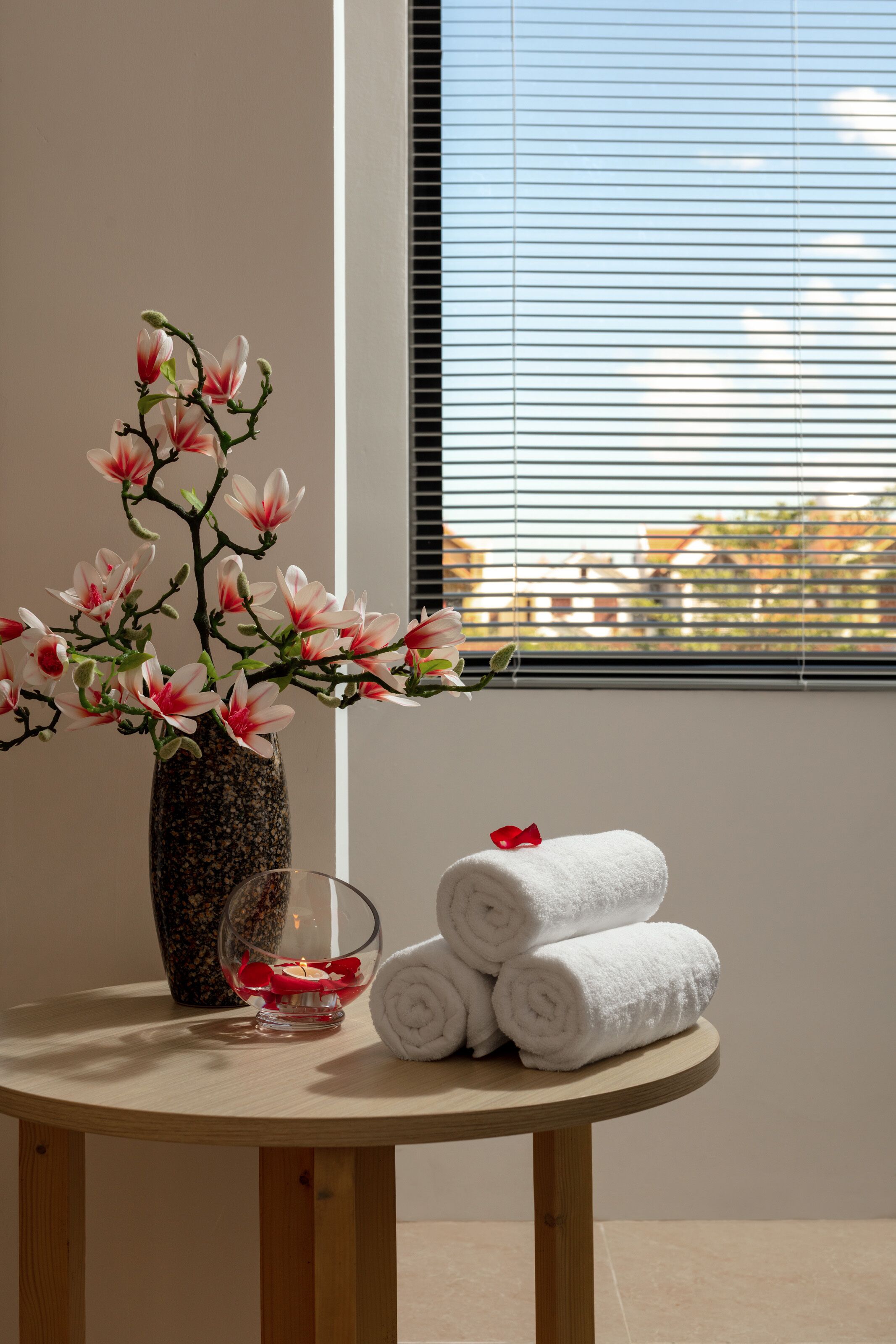 A round wooden table with a vase of pink flowers, a glass candle holder with a lit candle, and rolled white towels.