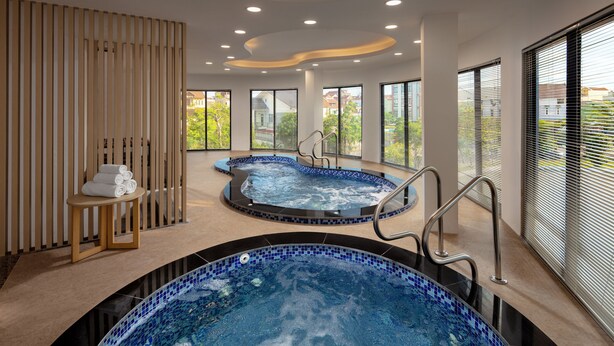 Area piscina interna moderna con due vasche idromassaggio, un pannello divisorio in legno e un tavolino con asciugamani.