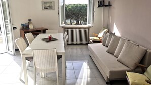 Un salón moderno con un sofá blanco, una mesa de comedor con sillas, un radiador y una ventana con vistas a árboles.