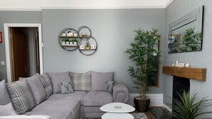 Un salon moderne avec un canapé d’angle gris, une petite table blanche, une grande plante en pot et une cheminée.