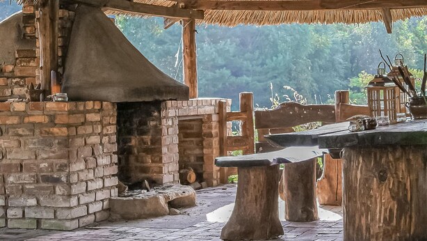 Ein Outdoor-Kochbereich mit einem Backofen aus Ziegeln, Holztischen und einem Reetdach.
