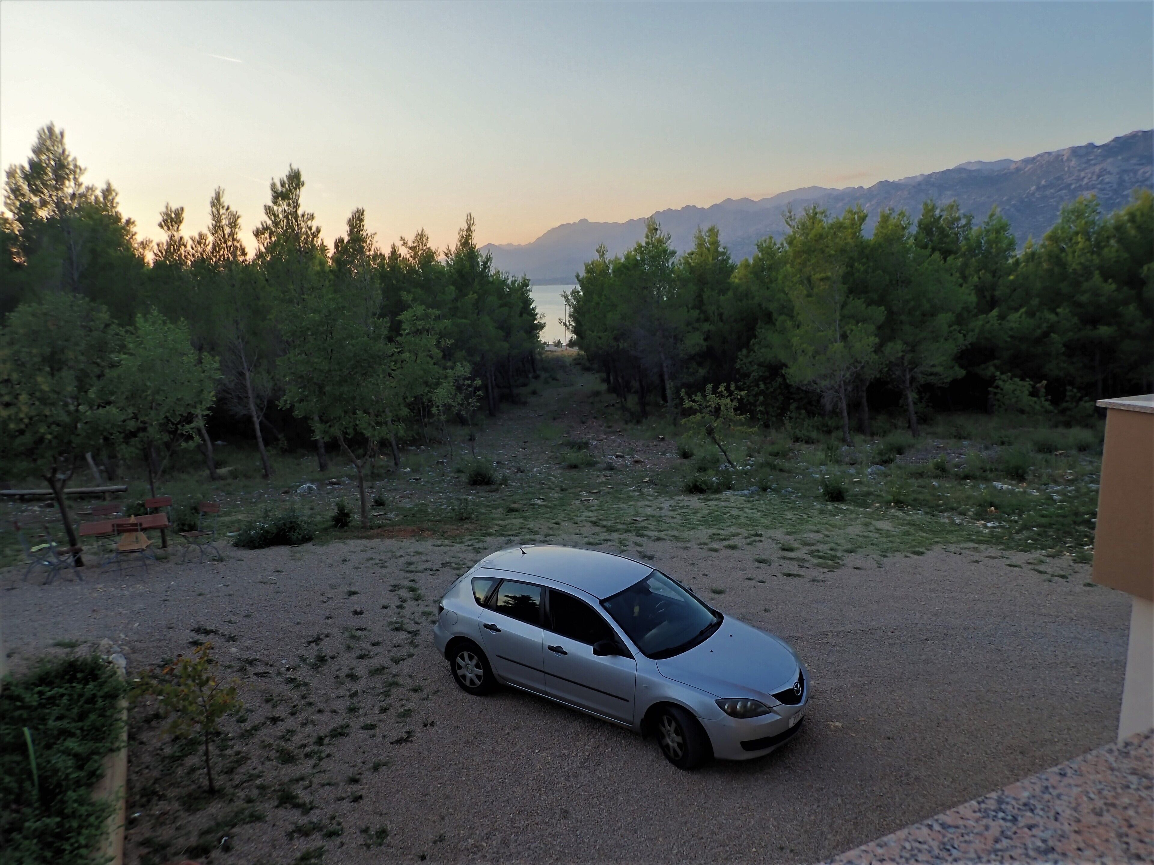 A silver car parked in a gravel area with trees and mountains in the background.
