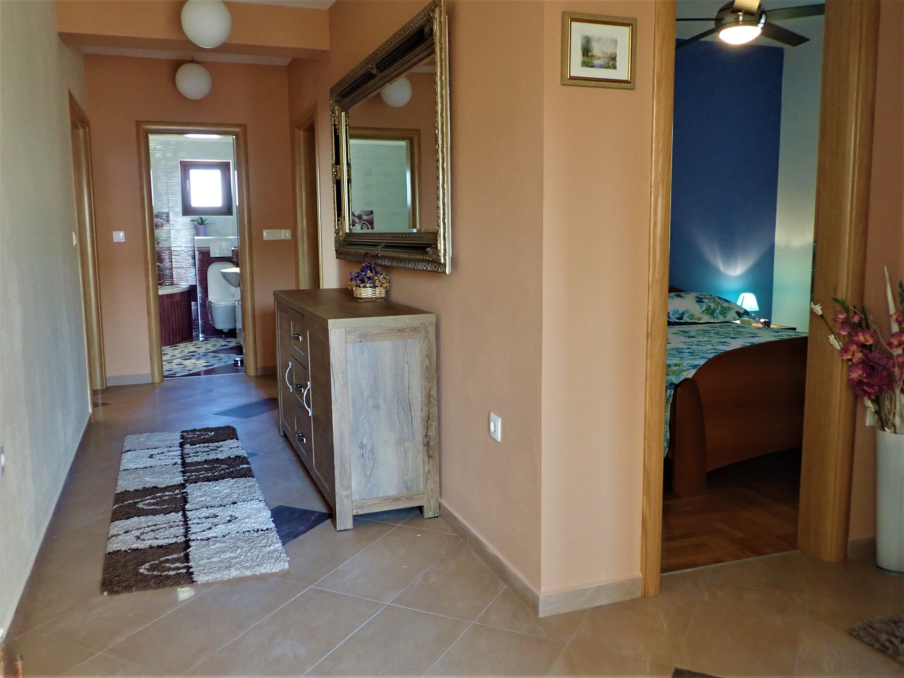 A hallway with a wooden dresser, a mirror, and a patterned rug. There is a bed with a floral bedspread in the background.