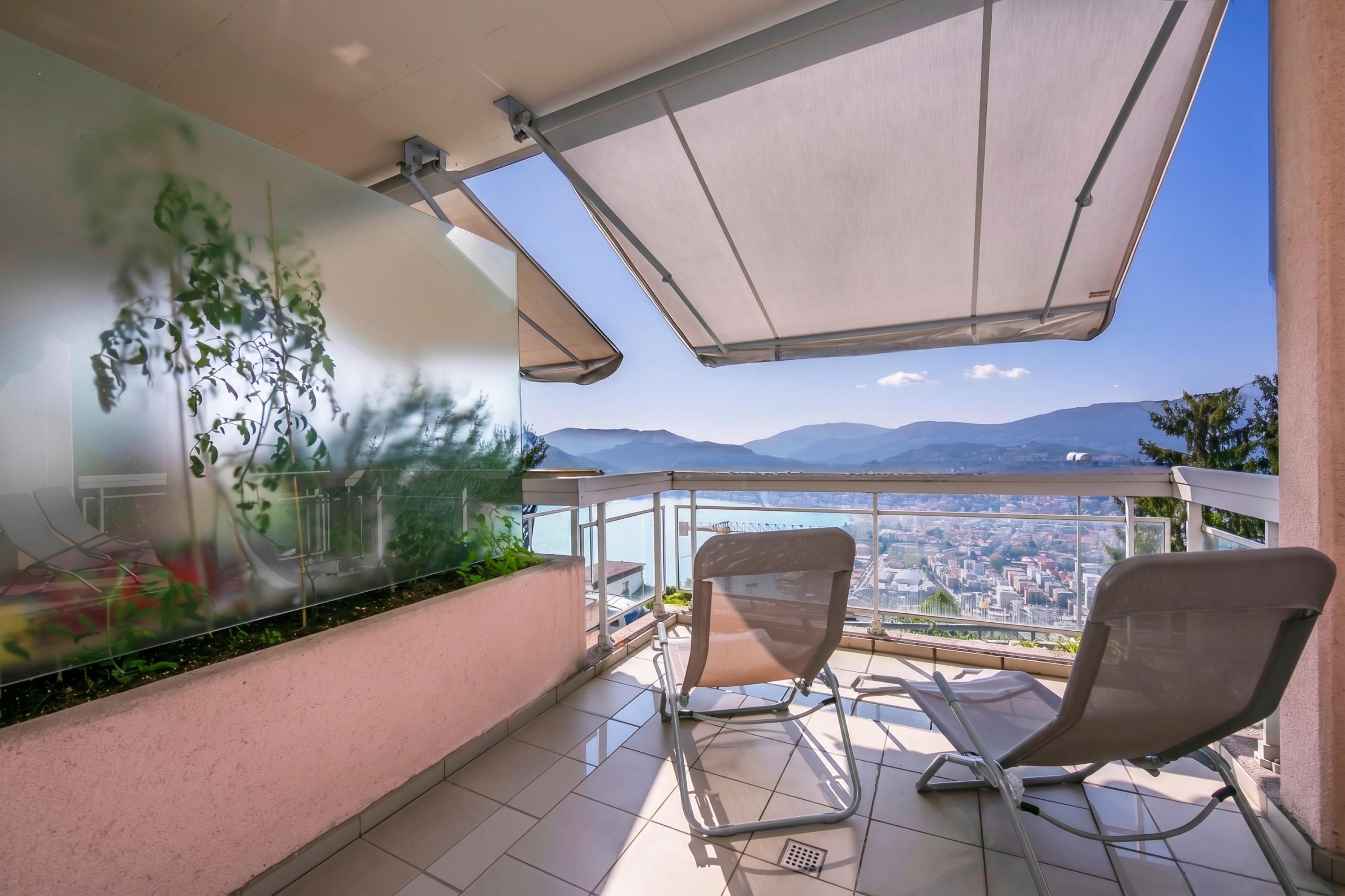 Un balcon avec deux chaises, une paroi en verre ornée de plantes et une vue sur la ville.