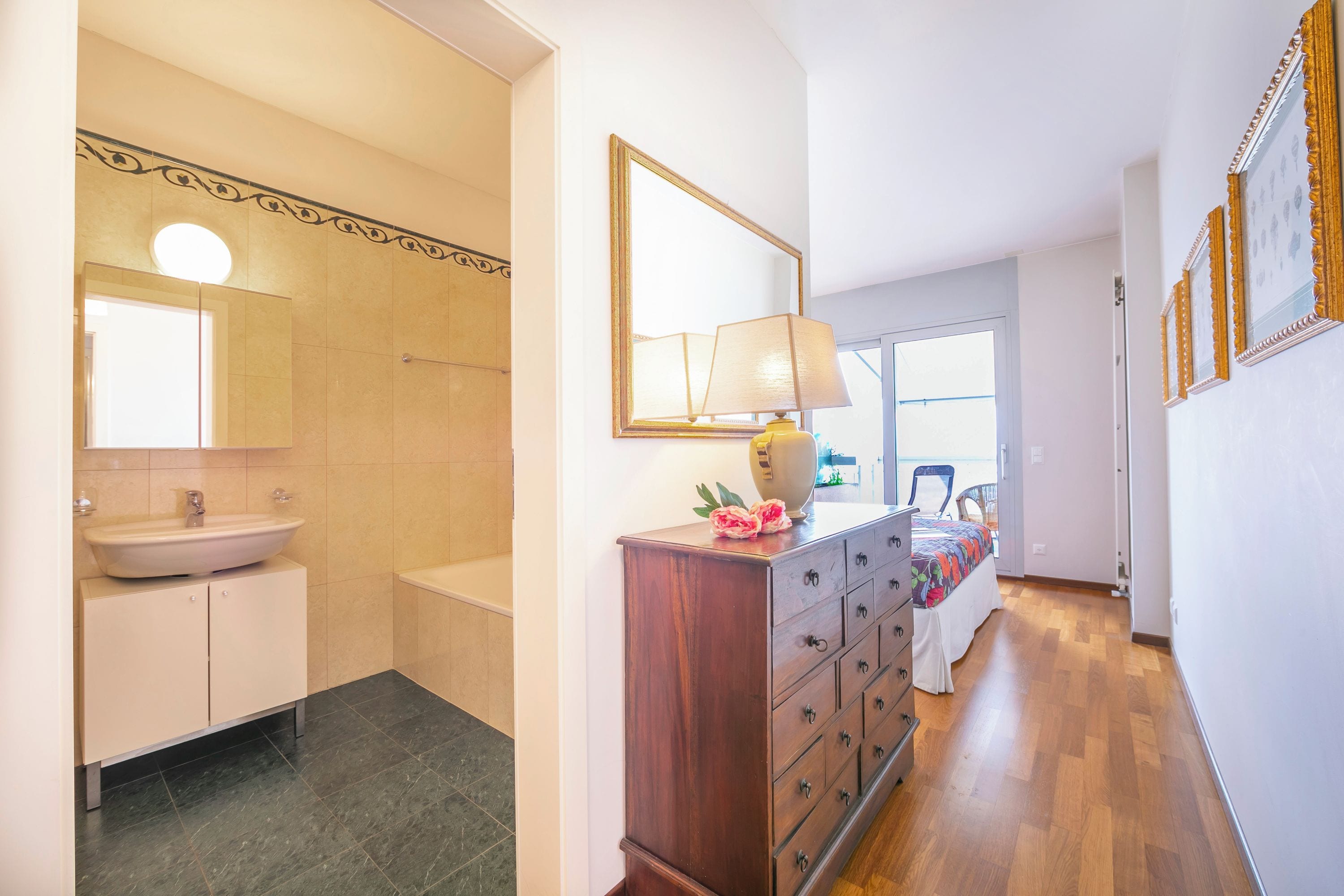 Un baño con lavamanos, espejo y tocador, un aparador de madera con cajones, y una cama con una colcha colorida.