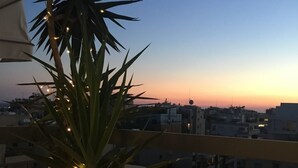 Un balcón con una planta en maceta y luces de cadena, con vista a un paisaje urbano al atardecer.