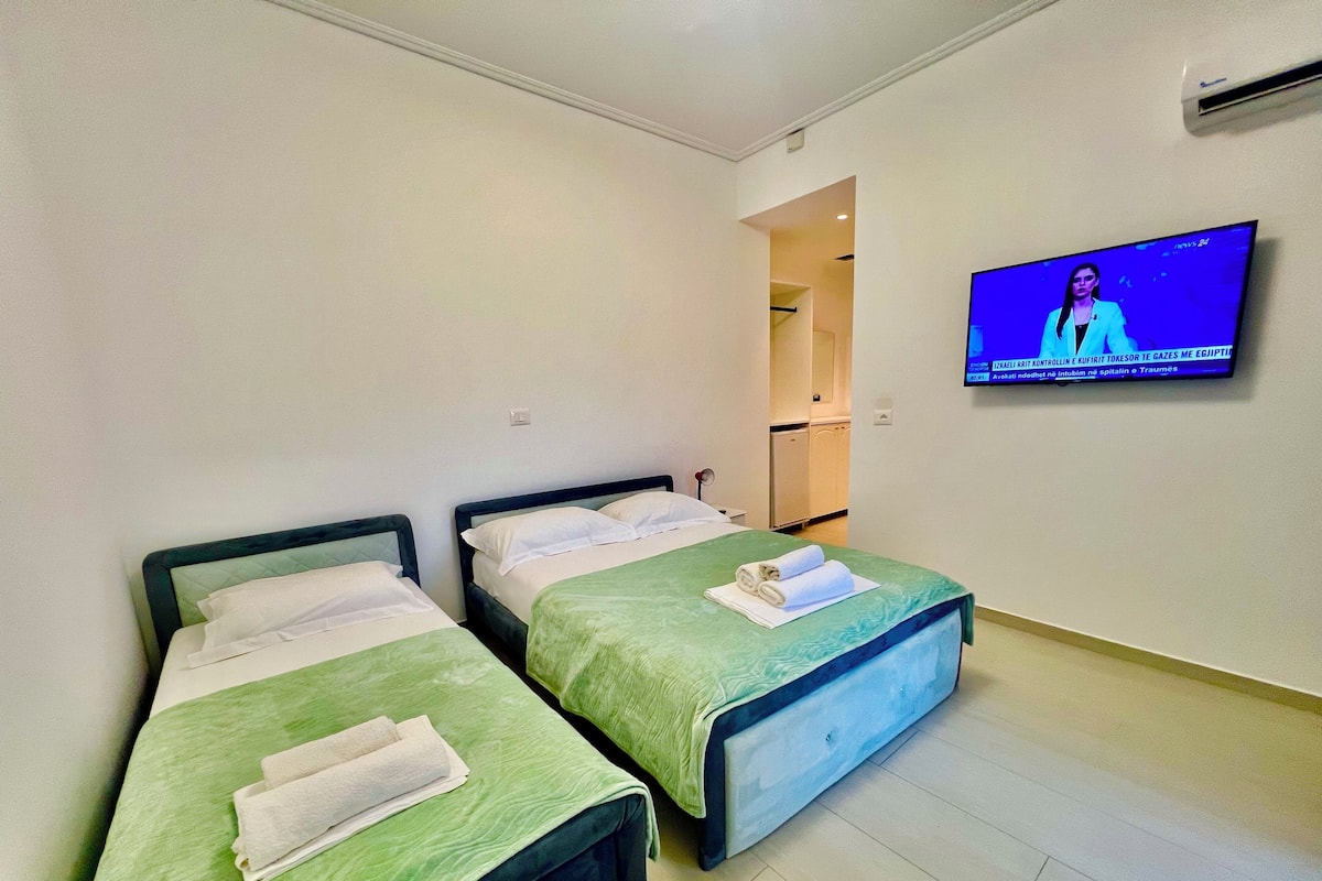 Une chambre d’hôtel avec deux lits simples, chacun recouvert d’une couverture verte et agrémenté d’oreillers blancs, et un téléviseur à écran plat fixé au mur.
