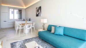 Un soggiorno moderno con un divano color petrolio, una zona pranzo con sedie bianche e una cucina con mobili bianchi.