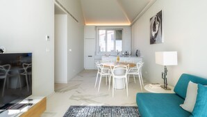 Un soggiorno moderno con una TV, un divano color petrolio, un'area pranzo con tavolo e sedie e una cucina sullo sfondo.