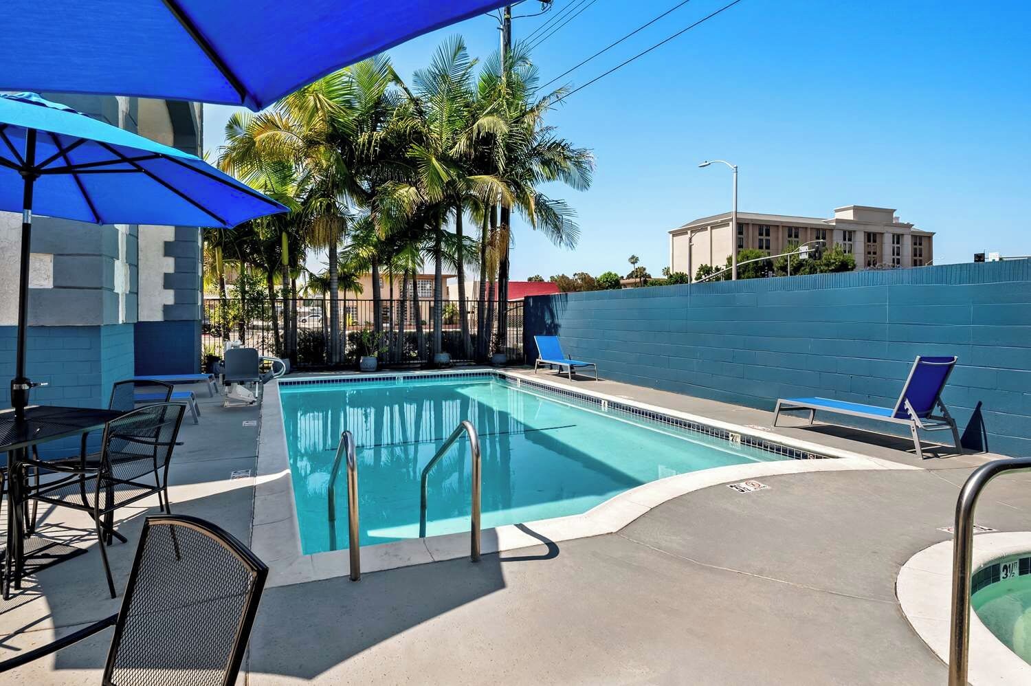Piscina com espreguiçadeiras, mesa e guarda-sóis, cercada por palmeiras e uma parede azul.