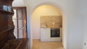 A kitchen with a tiled backsplash, a sink, and an oven. There is a wooden shelf with mugs and a door with a glass panel. - Il Trullo del Lauro (Laureto di Fasano)