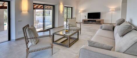 A spacious living area with a sofa, armchair, and coffee table. There is a TV and a lamp in the background.