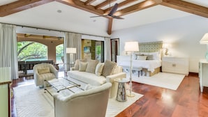 Un salon spacieux avec un plafond haut, des poutres en bois, un ventilateur de plafond, un ensemble de canapés, une table basse et une vue sur l’extérieur, accessible par une porte vitrée.