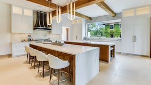 Une cuisine spacieuse dotée d’un grand îlot central, de poutres en bois et offrant une vue sur la verdure environnante.