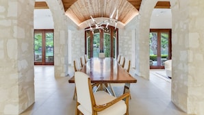 Un vaste espace dédié à la salle à manger, avec une longue table en bois et des chaises blanches à coussins, le tout entouré de murs en pierre et de grandes fenêtres offrant une vue sur la verdure.