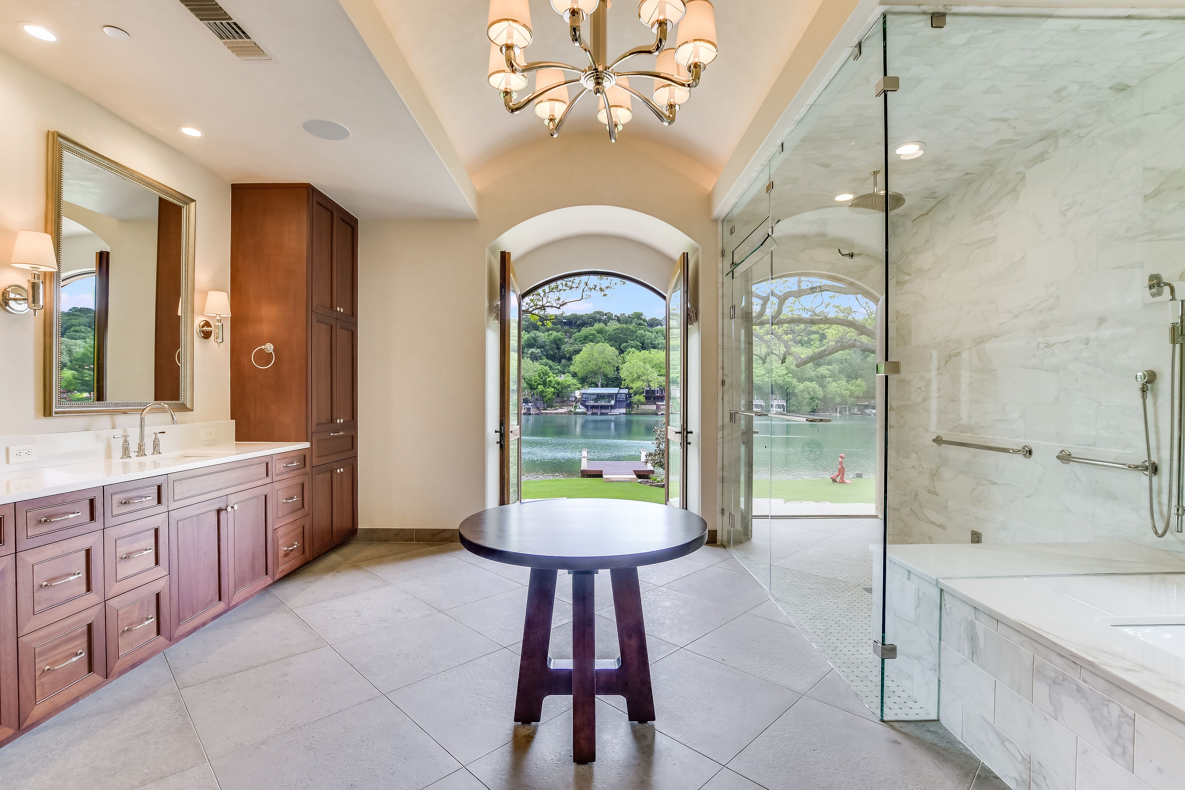 A modern bathroom with a marble vanity, a round stool, a glass-enclosed shower, and a view of a lake.