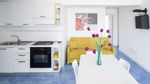 Une cuisine moderne avec des meubles blancs, une cuisinière et un évier. On y trouve un canapé jaune et une table à manger blanche, agrémentée d’un vase de fleurs violettes.