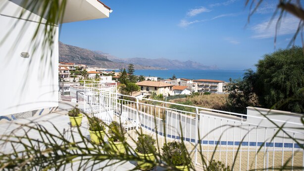 Un balcon agrémenté de plantes en pot, une balustrade blanche et une vue sur une ville côtière, avec des bâtiments et des montagnes en arrière-plan.