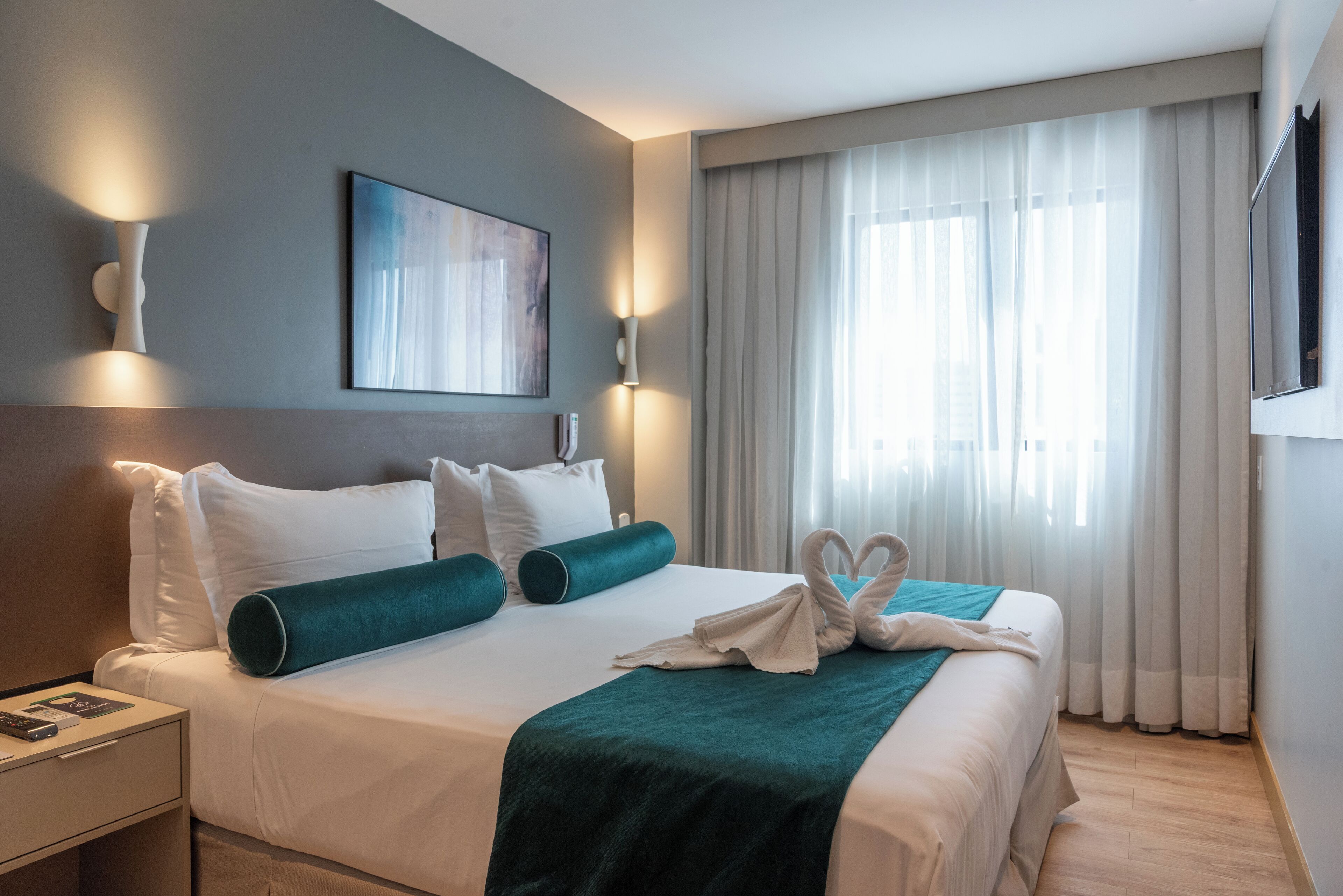 Una habitación de hotel con una cama bien hecha, una mesita de noche con lámpara, un cuadro en la pared y un amplio ventanal con cortinas.