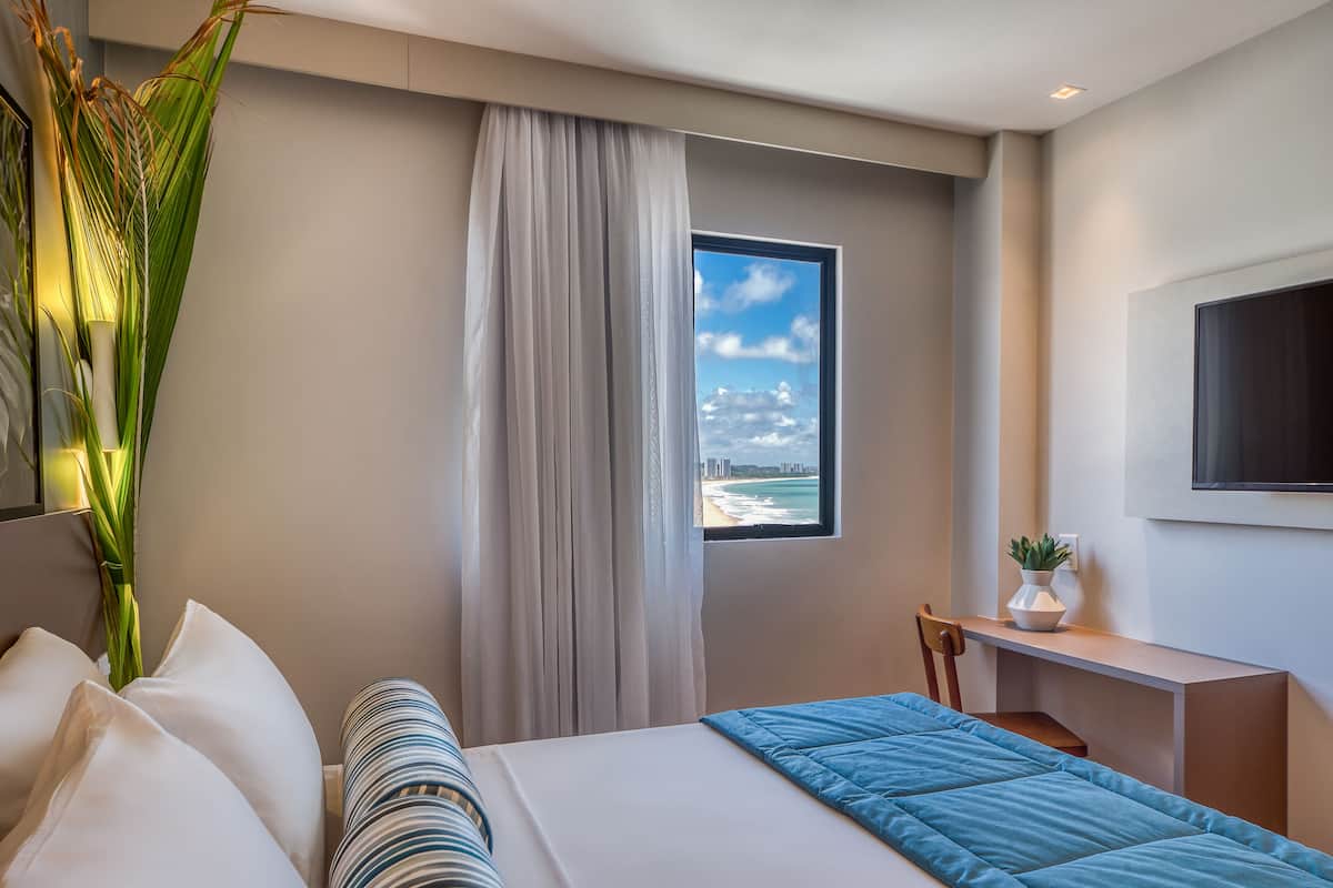 A modern hotel room with a bed, a desk, a chair, a window with a beach view, and a potted plant.