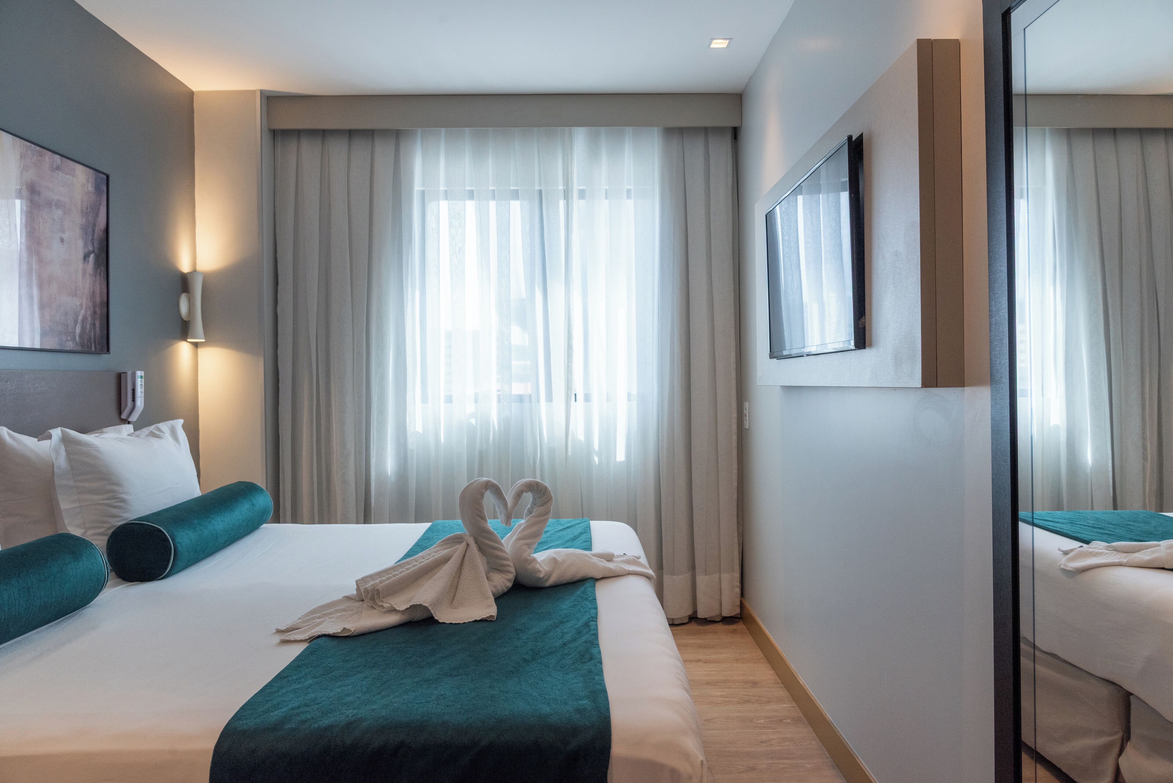 A hotel room with a neatly made bed, a large window with curtains, a wall-mounted TV, and a painting on the wall.