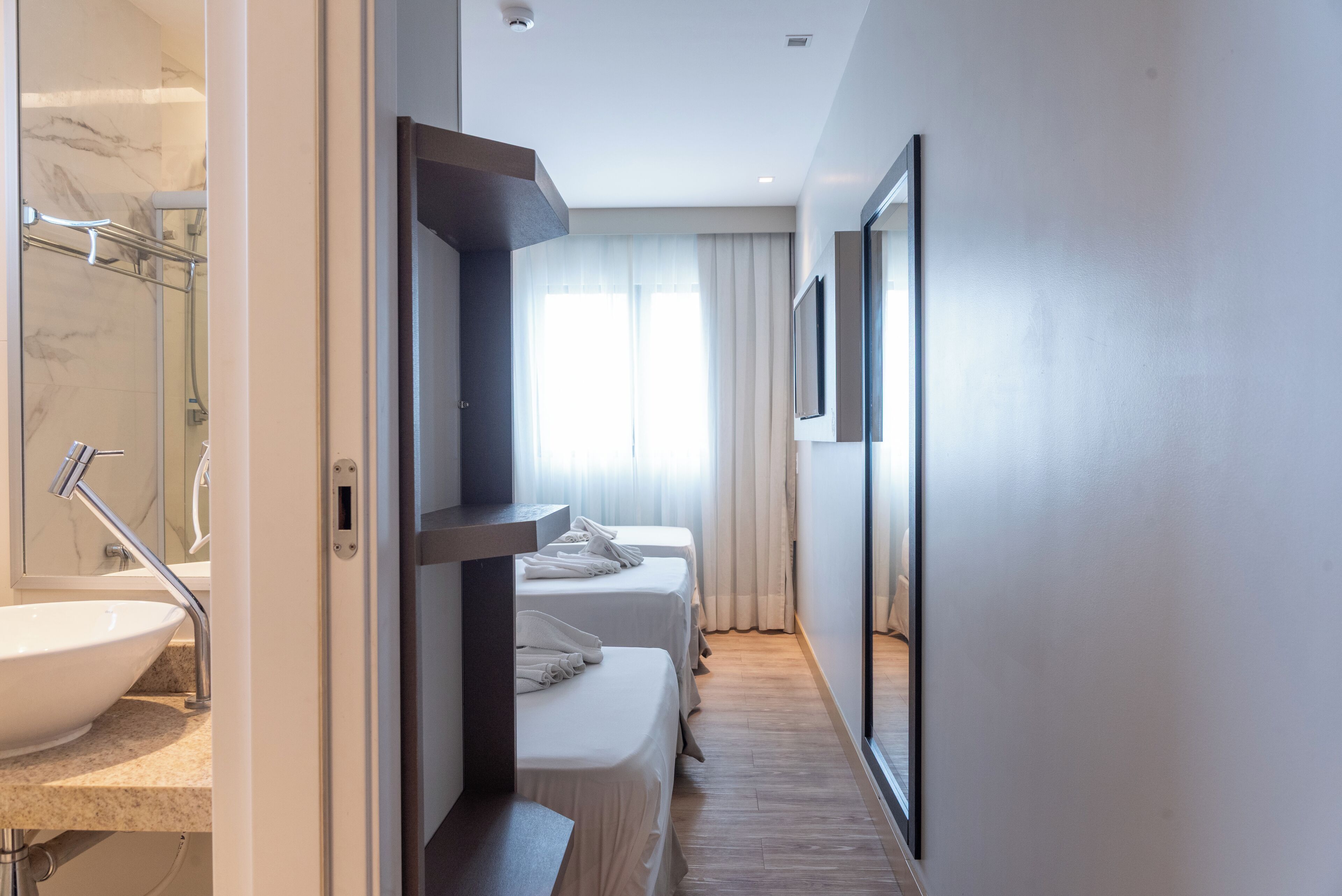 A modern hotel room with a large bathroom, two single beds, a large mirror, and a view of the city.