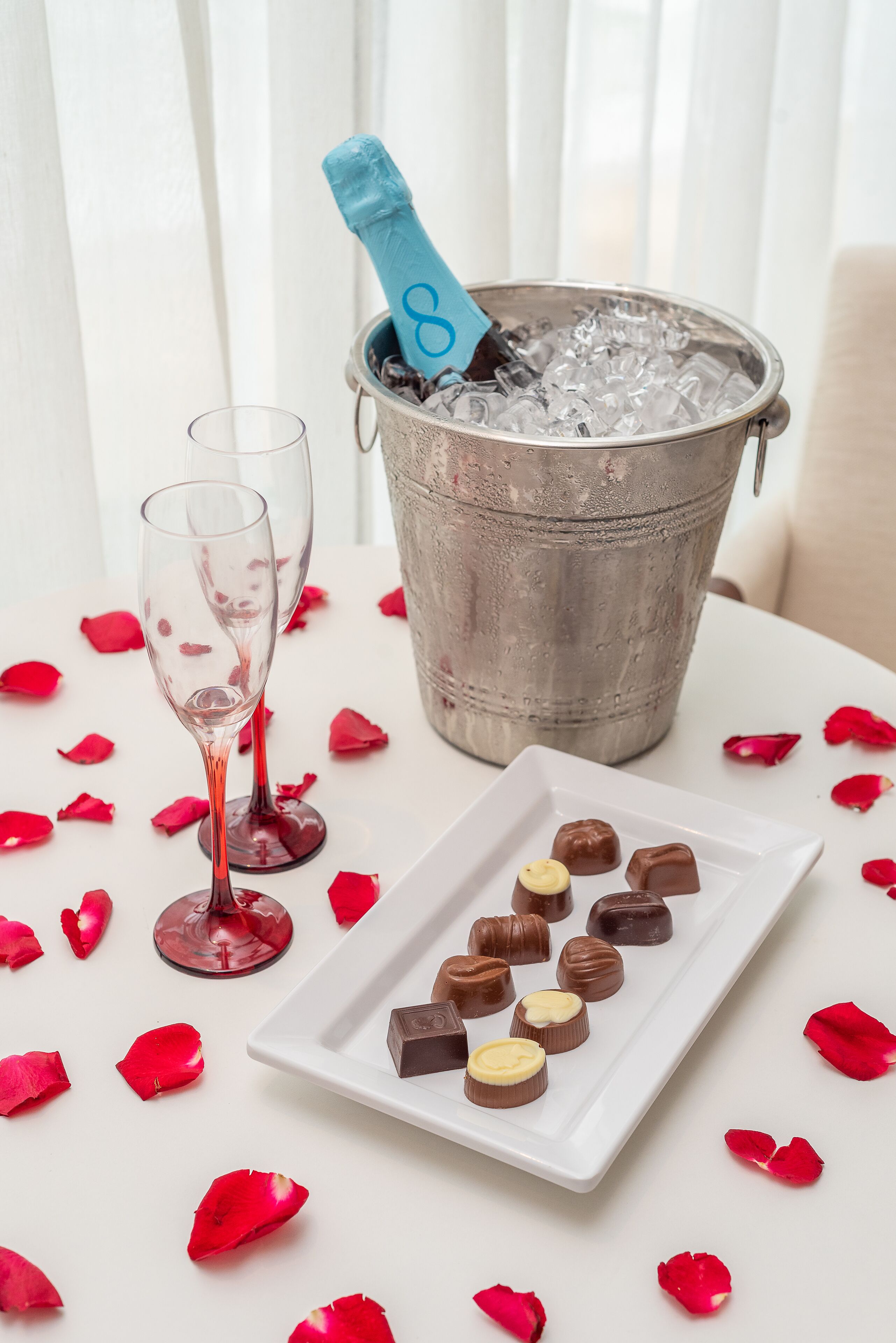 A table with a metal bucket containing a champagne bottle, two glasses, and a plate with assorted chocolates.