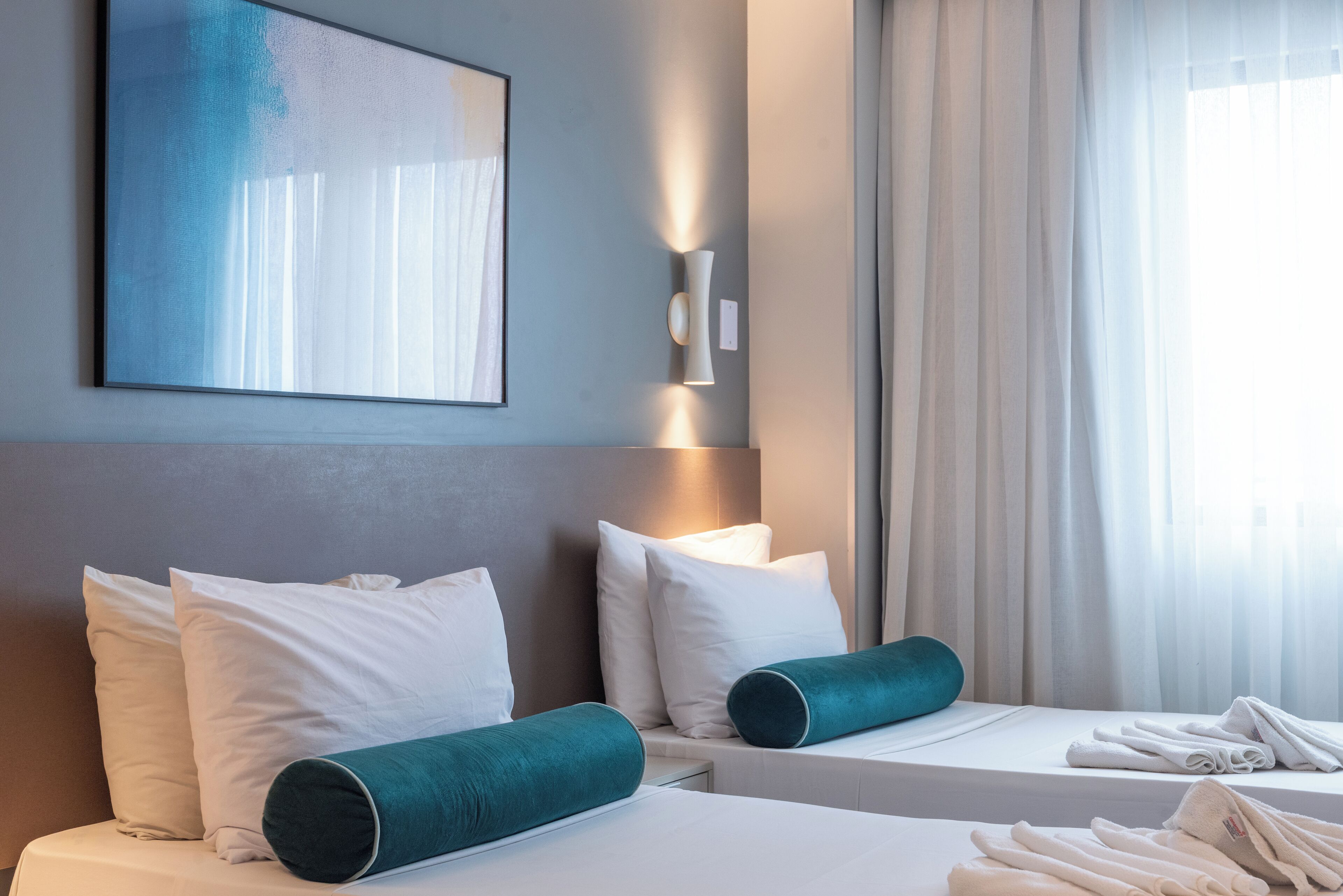 Quarto de hotel com duas camas, roupa de cama branca, almofadas verde-água, luminária fixada na parede e um grande quadro abstrato.