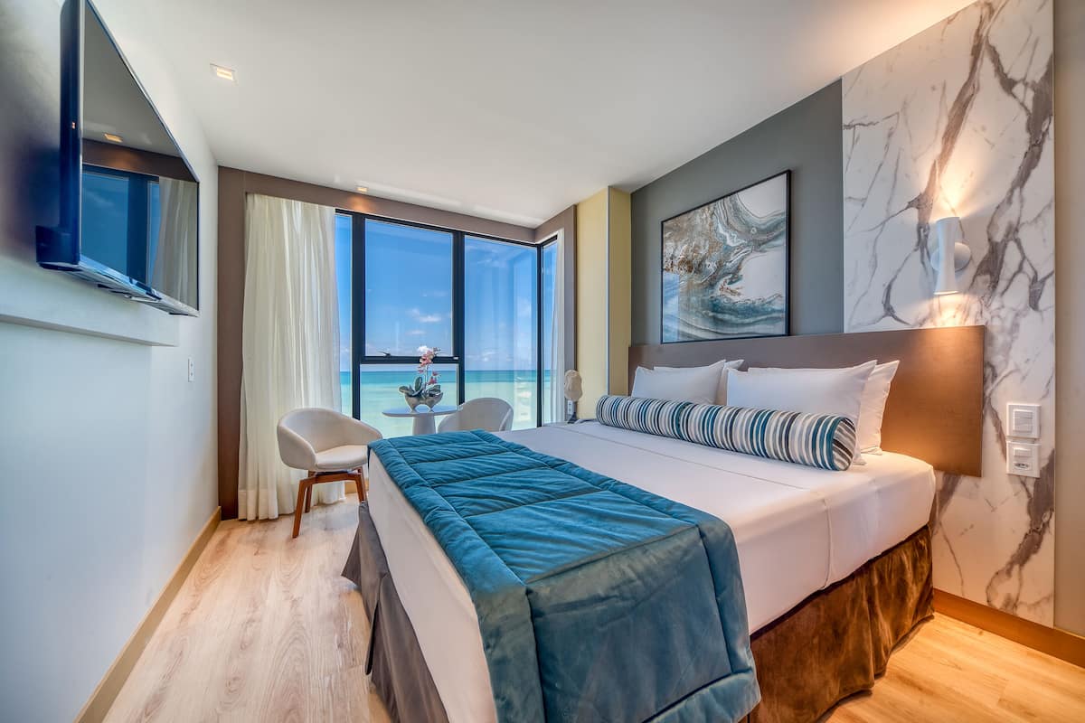 A modern hotel room with a large bed, a desk with a chair, a view of the ocean, and a marble wall.