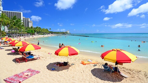Een strand met meerdere parasols, een zandstrand en helderblauw water.