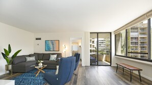 Een moderne woonkamer met een grijze bank, een blauwe fauteuil en een salontafel. Er is een groot raam met uitzicht op een balkon en andere gebouwen.