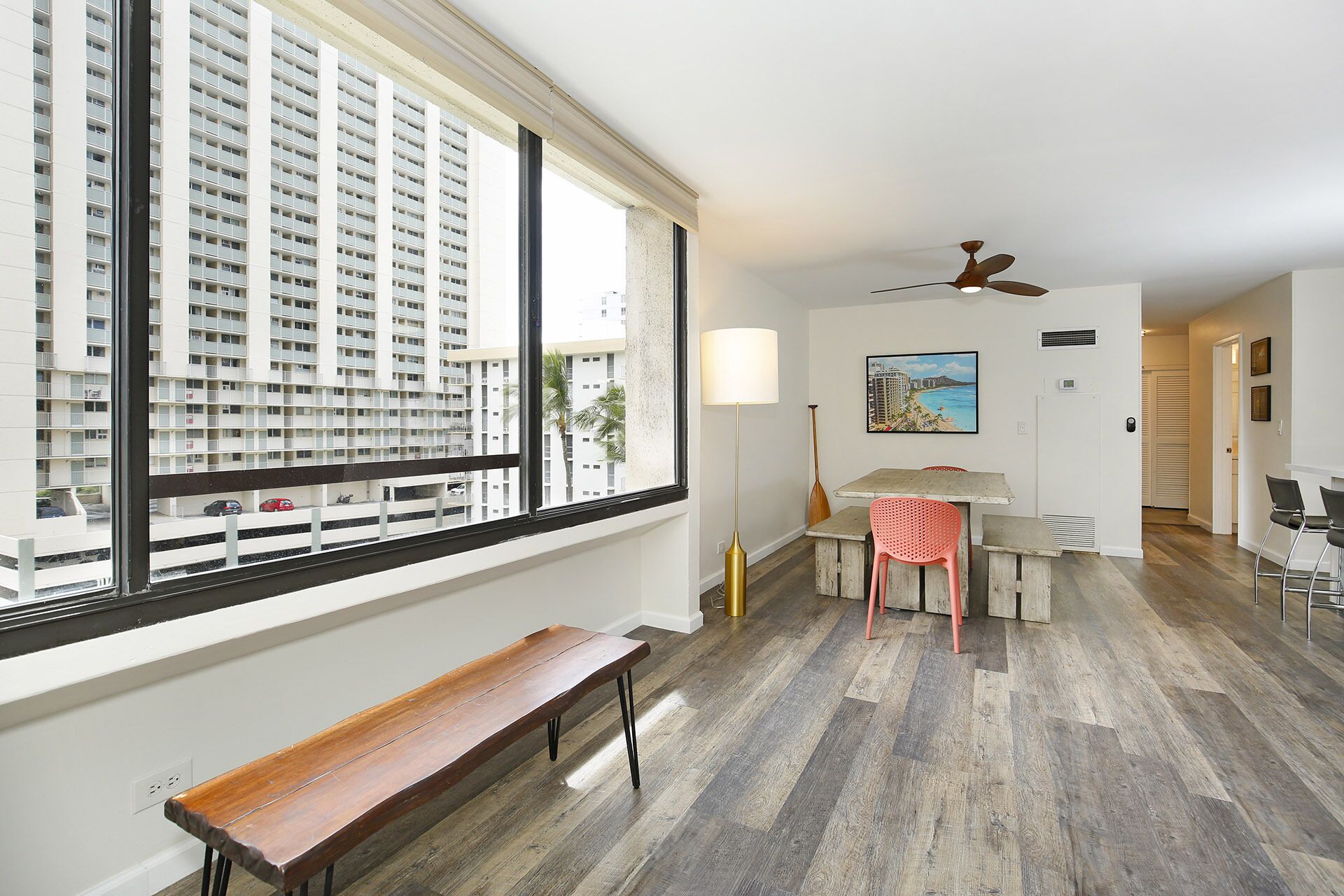 A modern apartment with a large window, a wooden bench, a dining area with a table and chairs, and a ceiling fan.