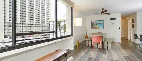 A modern apartment with a large window, a wooden bench, a dining area with a table and chairs, and a ceiling fan.