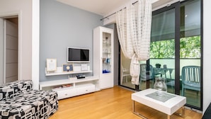Un salon moderne doté d’un téléviseur à écran plat, d’un meuble de rangement blanc, d’une table en verre avec des chaises et d’une porte-fenêtre coulissante donnant sur un balcon.