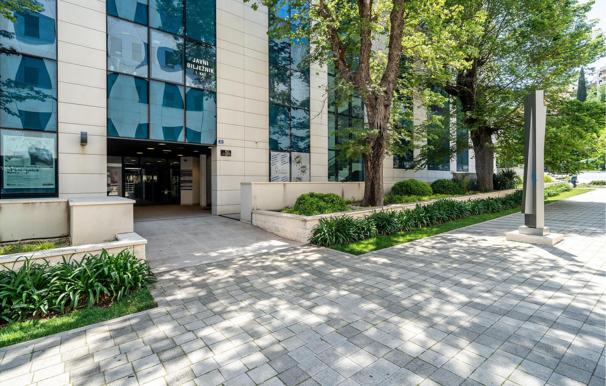 A modern building with a glass facade, a landscaped entrance area with trees and shrubs, and a paved walkway.