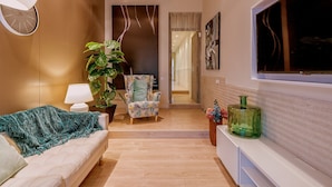 Una sala moderna con un sofá, un sillón estampado, una planta grande y un televisor sobre un mueble blanco.