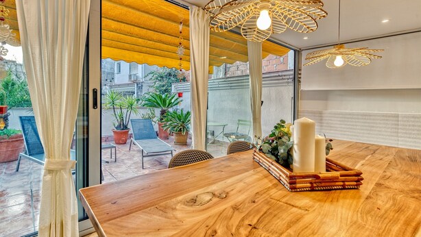 Una mesa de madera para comer con velas y una bandeja de frutas, un toldo amarillo y vista del patio exterior con plantas en macetas.