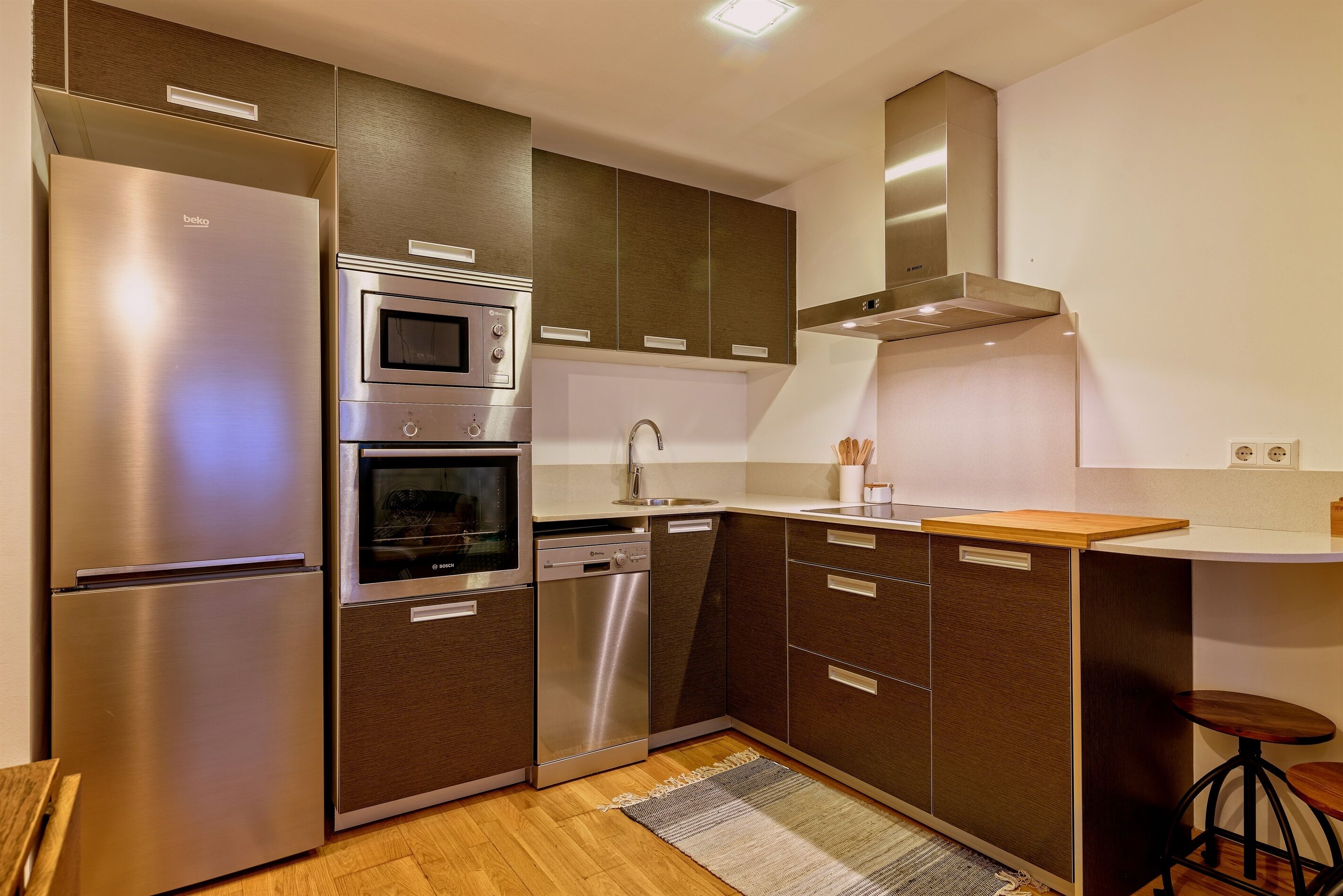 A compact kitchen with a built-in oven, microwave, and dishwasher, featuring dark wood cabinets and a stainless steel refrigerator.
