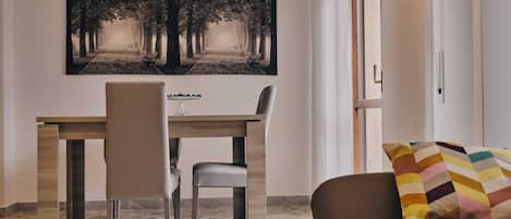 A dining area with a wooden table and chairs, a large black and white forest-themed painting on the wall, and a colorful cushion on a brown sofa.