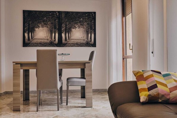 A dining area with a wooden table and chairs, a large black and white forest-themed painting on the wall, and a colorful cushion on a brown sofa.