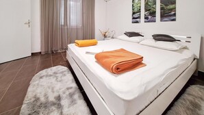 A neatly made bed with a folded orange towel, a white fluffy rug, and a window with a view of trees. - Amazing apartment in Mundanije (Mundanije)